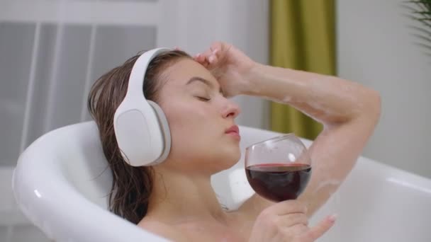 Mujer joven y relajada escuchando música en una burbuja. Primer plano de una joven relajada escuchando música con auriculares en un baño de burbujas y bebiendo vino tinto de una copa . — Vídeos de Stock