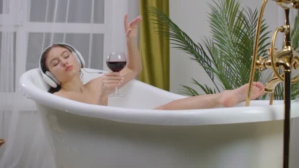 Primo piano di una giovane donna sdraiata in una vasca da bagno che ascolta musica con grandi cuffie in testa e beve vino rosso da un bicchiere. Concetto di relax e libertà — Video Stock