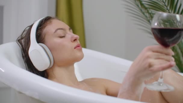 Hermosa joven disfrutando de un agradable baño con espuma, sosteniendo una copa de vino, Mujer escuchando música en el baño. Mujer escuchando música en baño de burbujas . — Vídeo de stock