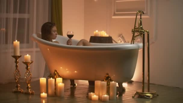 Une jeune belle brune caucasienne se trouve dans la salle de bain aux chandelles dans une agréable atmosphère de soirée reposant sur le stress et la détente en buvant du vin rouge dans un verre . — Video