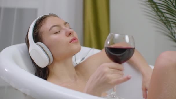 Hermosa joven disfrutando de un agradable baño con espuma, sosteniendo una copa de vino, Mujer escuchando música en el baño. Mujer escuchando música en baño de burbujas . — Vídeo de stock