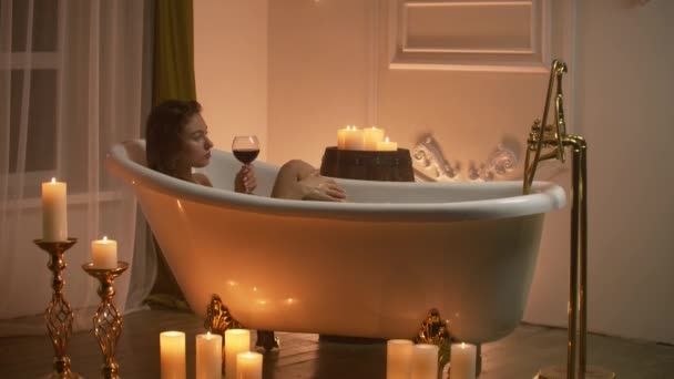 Женщина лежит в пенной ванне со свечами, чтобы расслабиться и выпить вина. Романтическая расслабляющая атмосфера ароматерапии. Ванная комната со свечами — стоковое видео