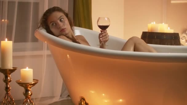 Jeune femme sexy avec corps nu droit et jambes propres couché et relaxant dans une baignoire en mousse blanche avec des bougies autour dans la salle de bain légère, boire de l'alcool à partir de verre de vin à l'intérieur . — Video