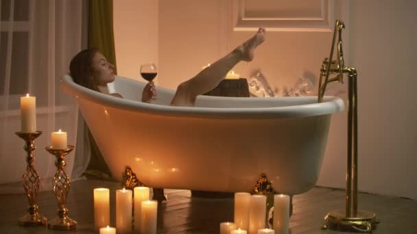 young sexy woman with straight naked body and clean legs lying and relaxing in white foam bath tub with candles around in light bathroom, drink alcohol from wine glass indoors.