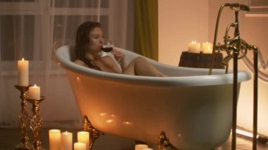 Kadın dinlenmek ve şarap içmek için mumların ışığıyla köpük banyosunda yatıyor. Romantik rahatlatıcı aromaterapi atmosferi. Mumlu banyo.