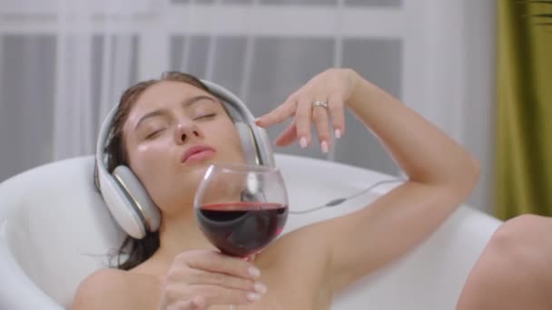 Mujer bastante joven escuchando música en los auriculares mientras toma el baño. Chica atractiva relajante tiene descanso en casa. Cuarto de baño ecológico interior. Aliviar el estrés y sentirse bien. Buen disfrute de contenido — Vídeo de stock