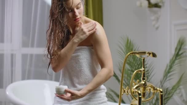 Женщина наносит крем на кожу в ванной комнате, сидя на ванне . — стоковое видео