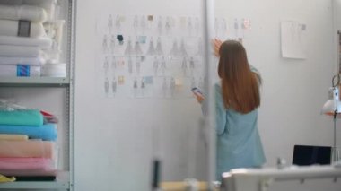 Kadın Moda Tasarımcısı. Masasının arkasındaki duvara iğnelenmiş çizimler ve çizimlere bakıyor. Stüdyo Sunny 'dir. Kişisel Bilgisayar, Renkli Kumaşlar, Dikiş Ögeleri Görünür