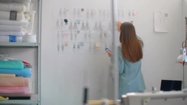 Diseñadora de moda femenina que mira dibujos y bocetos que se fijan a la pared detrás de su escritorio. El estudio es Sunny. Ordenador personal, telas coloridas, artículos de costura son visibles — Vídeo de stock