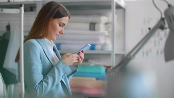 Een jonge vrouw in een designer tailoring Studio communiceert met klanten en bespreekt het gebruik van een mobiele telefoon en benadert een Board met schetsen op jurken — Stockvideo