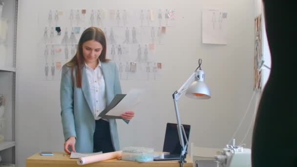 Female Fashion Designer Kijkend naar Tekeningen en Schetsen die zijn vastgepind aan de muur achter haar bureau. De studio is Sunny. Personal Computer, Kleurrijke stoffen, Naaiartikelen zijn zichtbaar — Stockvideo