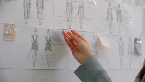 Modedesignerin beim Betrachten von Zeichnungen und Skizzen, die hinter ihrem Schreibtisch an die Wand geheftet sind. Studio ist sonnig. Personal Computer, bunte Stoffe, Nähartikel sind sichtbar — Stockvideo