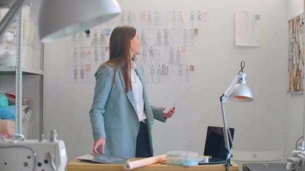 Joven diseñador de moda de pie con la espalda a la cámara mirando dibujos bocetos colgando en la pared. Mujer está pensando en la nueva colección de ropa de moda . — Vídeos de Stock