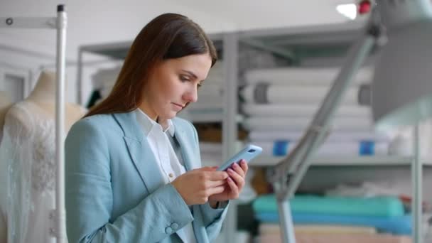 Una joven diseñadora con teléfono móvil en un taller de costura escribe mensajes a los clientes. Comunicación con los clientes pequeña empresa — Vídeos de Stock