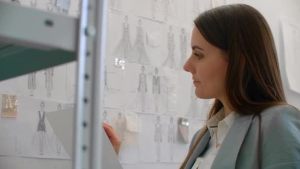 Female Fashion Designer Looking at Drawings and Sketches that are pinned to the Wall Behind Her Desk. Студия это Санни. Персональный компьютер, красочные ткани, швейные принадлежности являются видимыми — стоковое видео