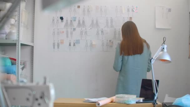 Jonge mode-ontwerper staat met rug naar camera te kijken naar tekeningen schetsen opknoping op de muur. Vrouw denkt na over nieuwe trendy kledingcollectie — Stockvideo