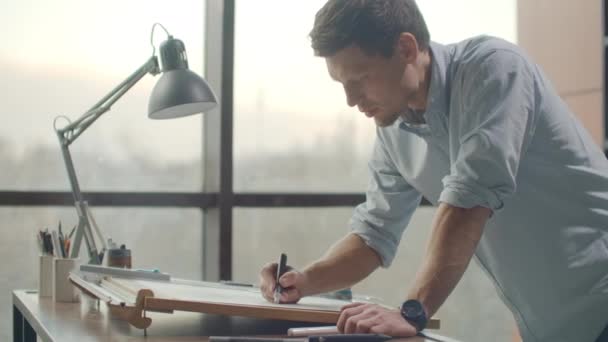 Um homem é um jovem arquiteto moderno sentado a uma mesa desenhando um plano do edifício e desenvolvendo um design, sentado em um escritório no loft Sitel perto de uma grande janela . — Vídeo de Stock