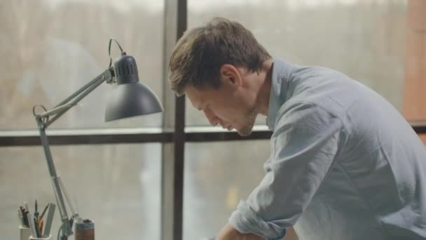 Ein Mann ist ein junger moderner Architekt, der an einem Tisch sitzt, einen Plan des Gebäudes entwirft und einen Entwurf entwickelt, in einem Büro im sitel Loft neben einem großen Fenster. — Stockvideo