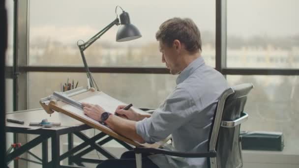 Un architecte masculin assis à une table dessine un plan du bâtiment et est engagé dans le développement du design, assis dans le bureau dans le grenier Sitel près d'une grande fenêtre — Video