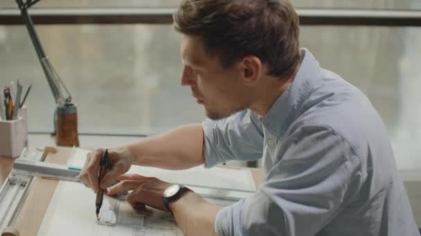Un arquitecto masculino sentado en una mesa dibuja un plano del edificio y se dedica al desarrollo de diseño, sentado en la oficina en el loft Sitel cerca de una gran ventana — Vídeos de Stock