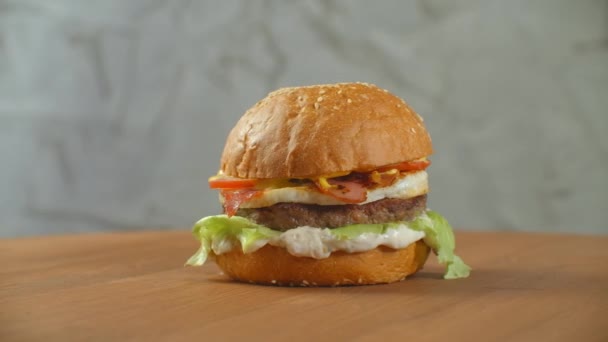 Hamburger se točí na dřevěné desce. Burger se salátovým sýrem a rajčaty se otáčí proti šedé stěně. — Stock video