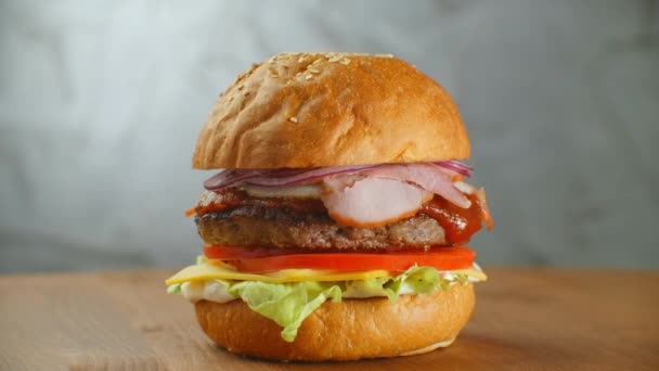 Гамбургер обертається на дерев'яній дошці. Бургери з котлетиним салатом з сиром і помідорами обертаються навколо сірої стіни . — стокове відео