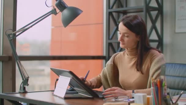 Ένας επαγγελματίας σχεδιαστής στο γραφείο σχεδιάζει με μια γραφίδα σε ένα tablet που κάθεται σε ένα γραφείο με τεράστια παράθυρα σε στυλ loft. Σύγχρονο γραφείο του γραφίστα και δημιουργού — Αρχείο Βίντεο