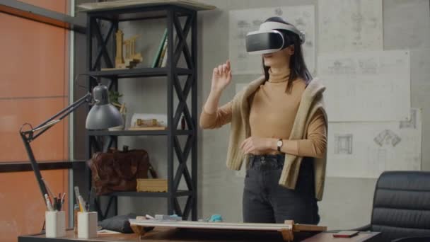 Молода жінка-інженер в офісі в шоломі віртуальної реальності з руками робить рухи, що імітують роботу графічного інтерфейсу — стокове відео