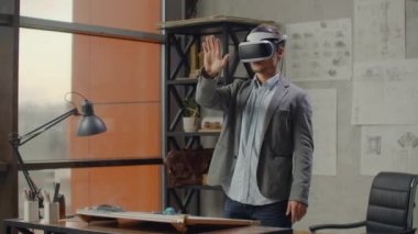 Ofisteki büyük pencereli modern mimar sanal gerçeklik miğferinin içinde duruyor. Ofisten çıkmadan projeyi yönetmek için jestler yapıyor. İnşaat kontrolü. Tasarım projesi.