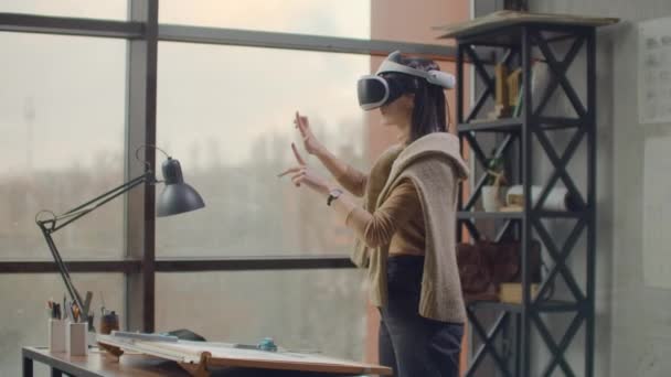 Femme architecte moderne dans le bureau avec de grandes fenêtres se dresse dans un casque de réalité virtuelle utilise des gestes pour gérer le projet sans quitter le bureau. Contrôle de construction. Projet de conception . — Video