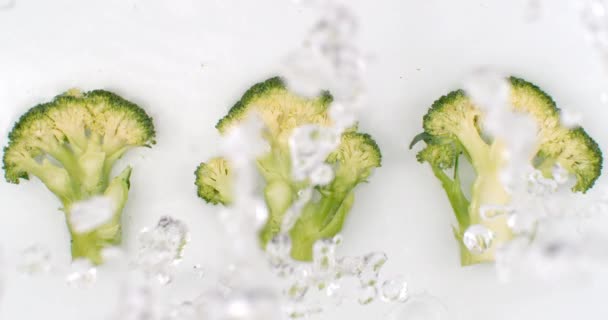 L'acqua schizza al rallentatore. Vista dall'alto: tre pezzi di broccoli verdi lavati con acqua su fondo bianco . — Video Stock