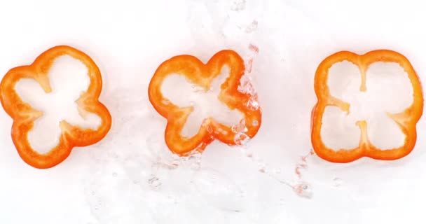 Ovanifrån: tre skivor röd paprika tvättas med vatten på en vit bakgrund. Vattenstänk i slow motion — Stockvideo