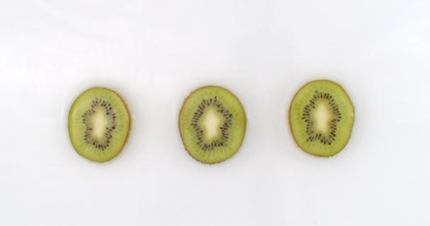 Vattenstänk i slow motion. Ovanifrån: tre bitar kiwi tvättas med vatten på en vit bakgrund. — Stockvideo