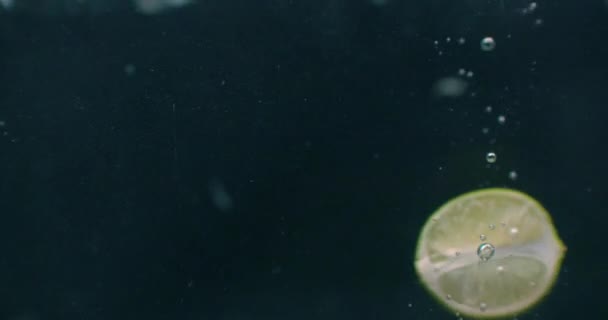 Les tranches de chaux tombent et flottent dans l'eau, fond noir, au ralenti. Des tranches de citron vert tombent et flottent dans l'eau, fond noir, ralenti .
