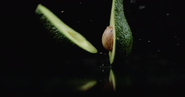 Eine reife geschnittene grüne Avocado fällt auf ein Glas mit Wasserspritzern in Zeitlupe auf dunklem Hintergrund. — Stockvideo