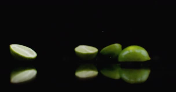 Several green ripe limes fall on the glass with splashes of water in slow motion on a dark background. — Stock Video