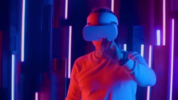 Uomo senza volto con auricolare VR nello spazio buio con lampade al neon, utente che gira la testa da un lato all'altro guardando la realtà virtuale, sparando attraverso razzi colorati e bokeh in primo piano . — Video Stock