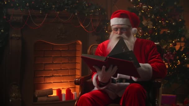 充满欢乐的正宗圣塔条款翻阅着红皮书，背景上有壁炉和圣诞树- -圣诞精神的概念近在咫尺 — 图库视频影像