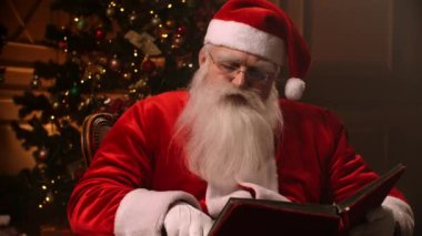 Santa Claus with glasses and a white beard, reading a red book on the background of a Christmas tree and a fireplace.