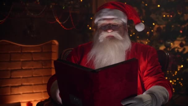 赤い表紙の本のページをめくる喜びの本格的なサンタの句、暖炉とクリスマスツリーを背景に-クリスマスの精神の概念を閉じる — ストック動画