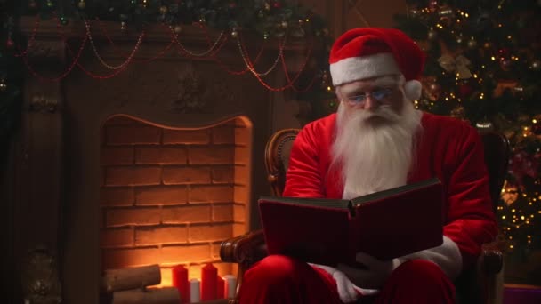 Santa Claus sedí ve speciálně zdobené místnosti, čte kouzelnou zářivou knihu - svátky a oslavy, vánoční duch koncept. — Stock video