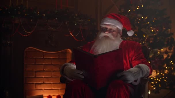 Realistic santa clause sitting near fireplace and christmas tree, opening a large magical book with shining pages - cristmas spirit, magic, dream concept. — Stok video