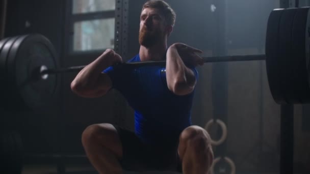 Slow motion: Ung atletisk man knäböjer med skivstång i gymmet. Mannen testar styrka genom att hålla en skivstång med tunga vikter på axlarna när han knäböjer. — Stockvideo