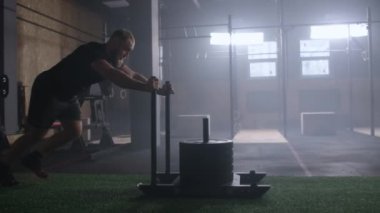 Willpower and motivation, camera follows young athletic man pushing heavy training sled training in gym slow motion