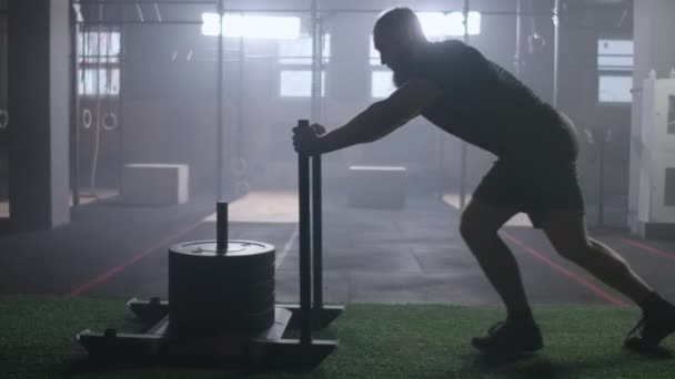 Concepto de motivación, joven atlético hombre rubio empujando pesado trineo de entrenamiento hacia el entrenamiento con cámara en el gimnasio cámara lenta — Vídeos de Stock