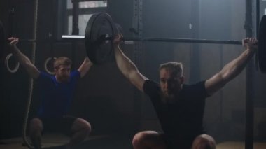 Two strong man doing weightlifting training together at the gym. Medium shot slow motion pan men exercising in gym with exercise equipments: Overhead squats, plate lunges, medicine ball, olympic rings