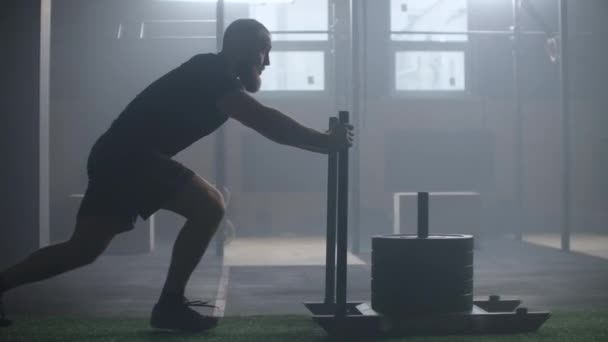 Joven hombre atlético caucásico fuerte que hace ejercicio con el trineo de entrenamiento de peso pesado hacia la cámara en el gimnasio grande cámara lenta . — Vídeo de stock