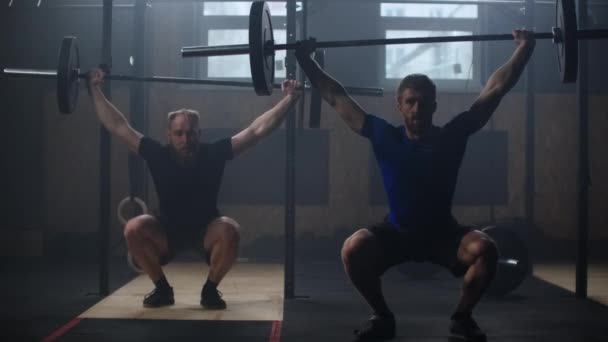 Deux hommes forts qui font de l'haltérophilie ensemble au gymnase. Poêle à mouvement lent de tir moyen hommes exerçant dans la salle de gym avec des équipements d'exercice : squats aériens, fentes de plaque, boule de médecine, anneaux olympiques — Video