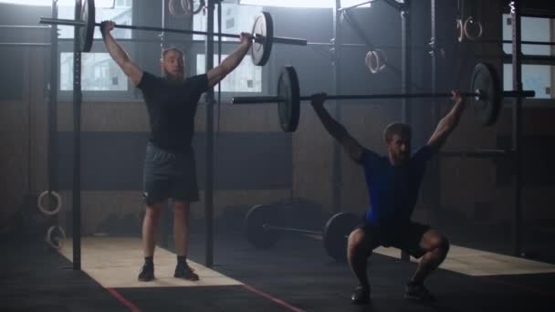 Dos hombres atléticos, atletas, haciendo ejercicios con la barra. En cámara lenta. Dos hombres fuertes haciendo levantamiento de pesas con barra en el gimnasio simultáneamente . — Vídeo de stock