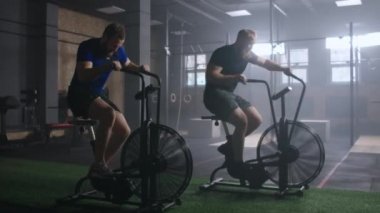 2 Young active man spinning air bike in gym. Male training on air bike in slow motion. Medium shot of fitness men working out with copy space.
